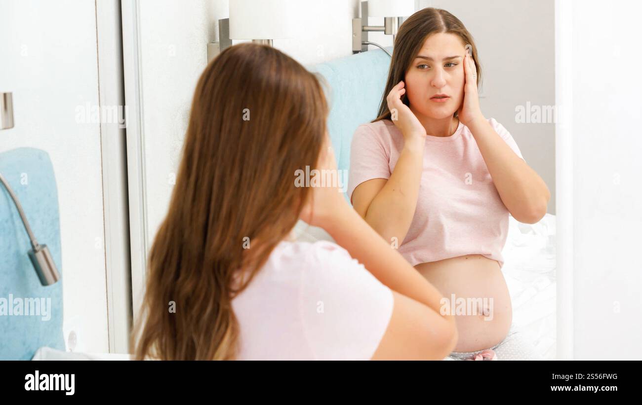 Portrait of pregnant woman suffering from head ache looking in her reflection in mirror. Concept of pregnancy healthcare and medical examination Stock Photo