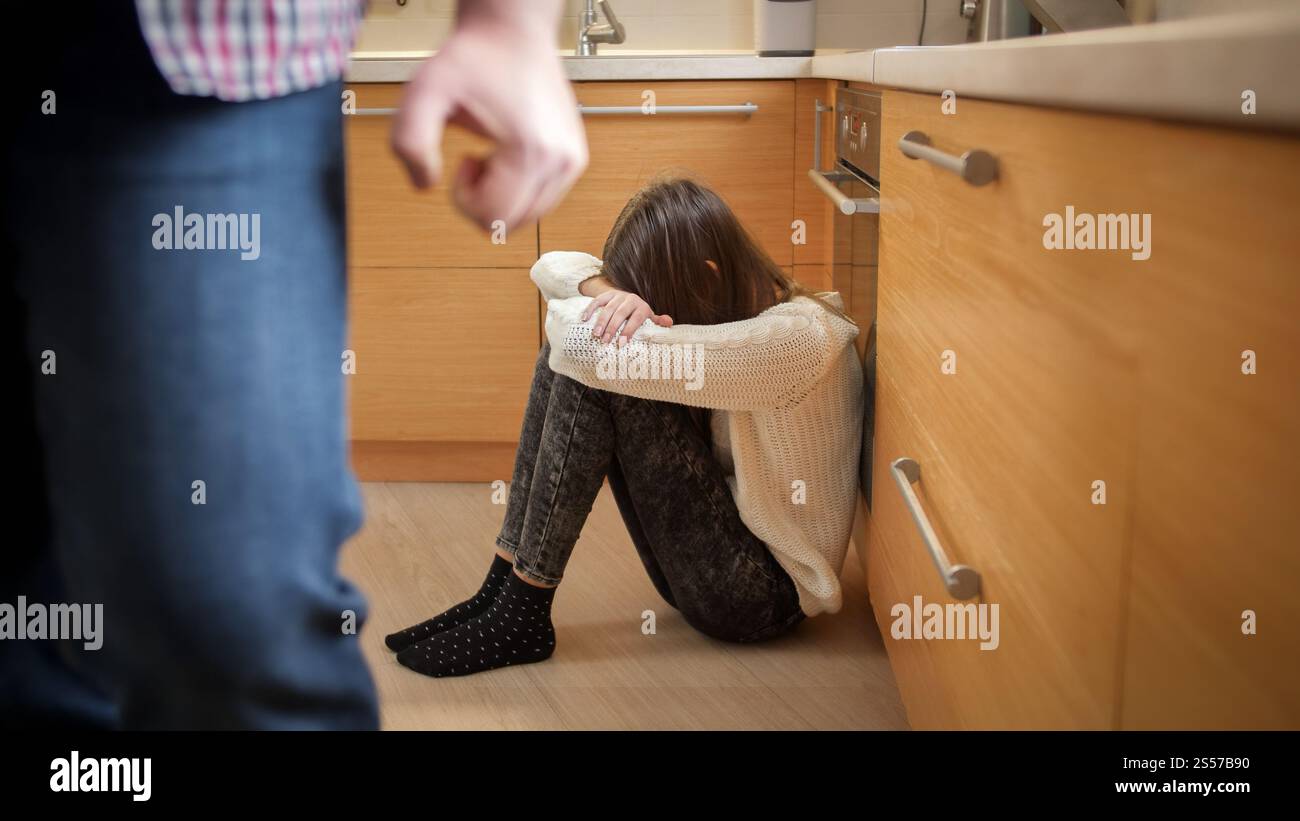 Angry father with clenched fist walking away from crying daughter sitting on kitchen floor. Concept of domestic violence and family aggression Stock Photo