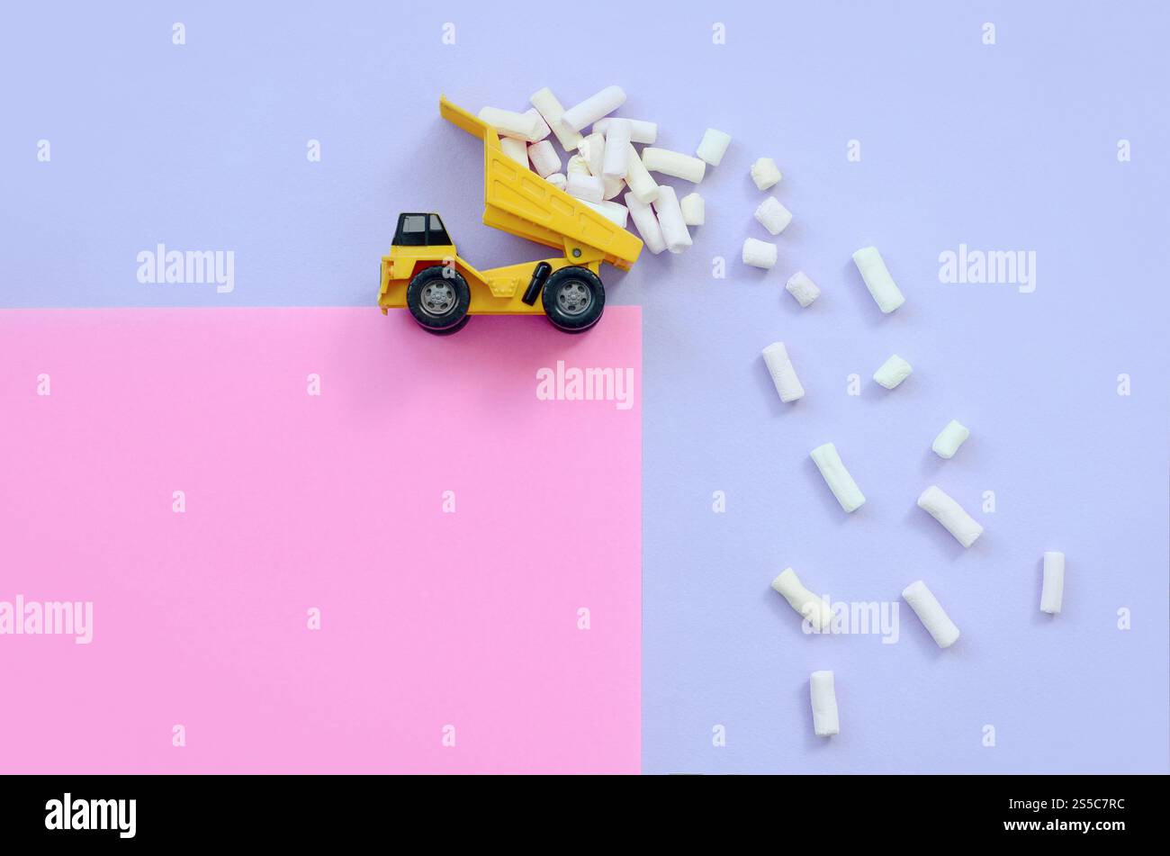 Yellow little toy dump truck throws marshmallow pieces from its raised back on a pastel violet and pink background. Flat lay minimal top view. Dump Stock Photo