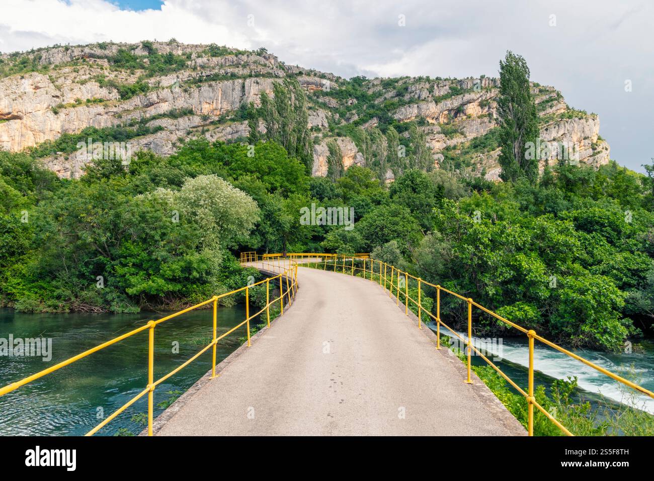 Yellow railed bridge over a river with lush greenery and towering cliffs in the background under a cloudy sky, Krka national park Stock Photo