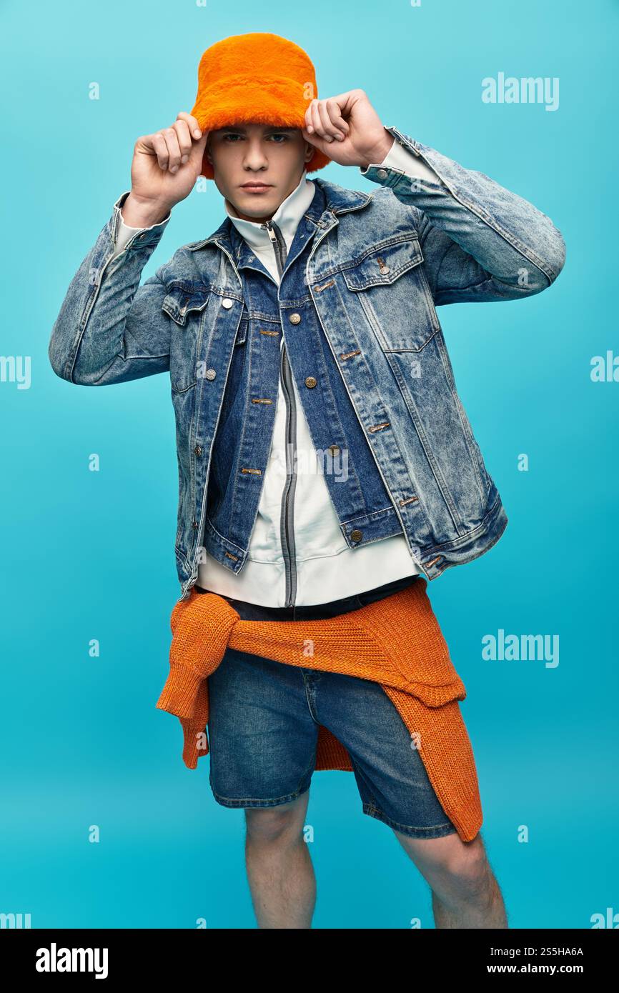 Young man in a denim jacket and bright orange hat poses against a blue ...