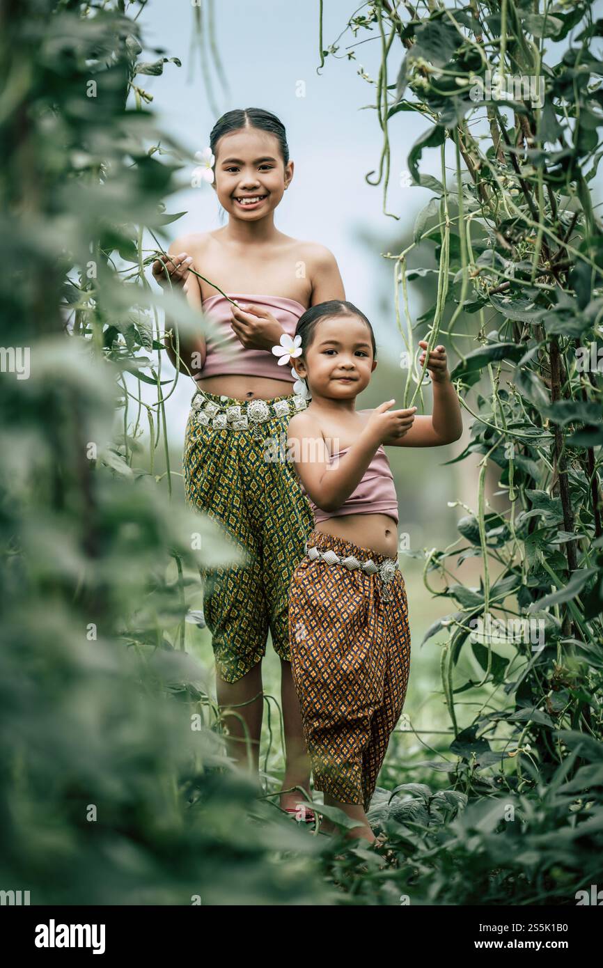 Portrait of two girls in Thai traditional dress and put white flower on her ear, standing in plant yard of long beans, They are smile with happiness Stock Photo