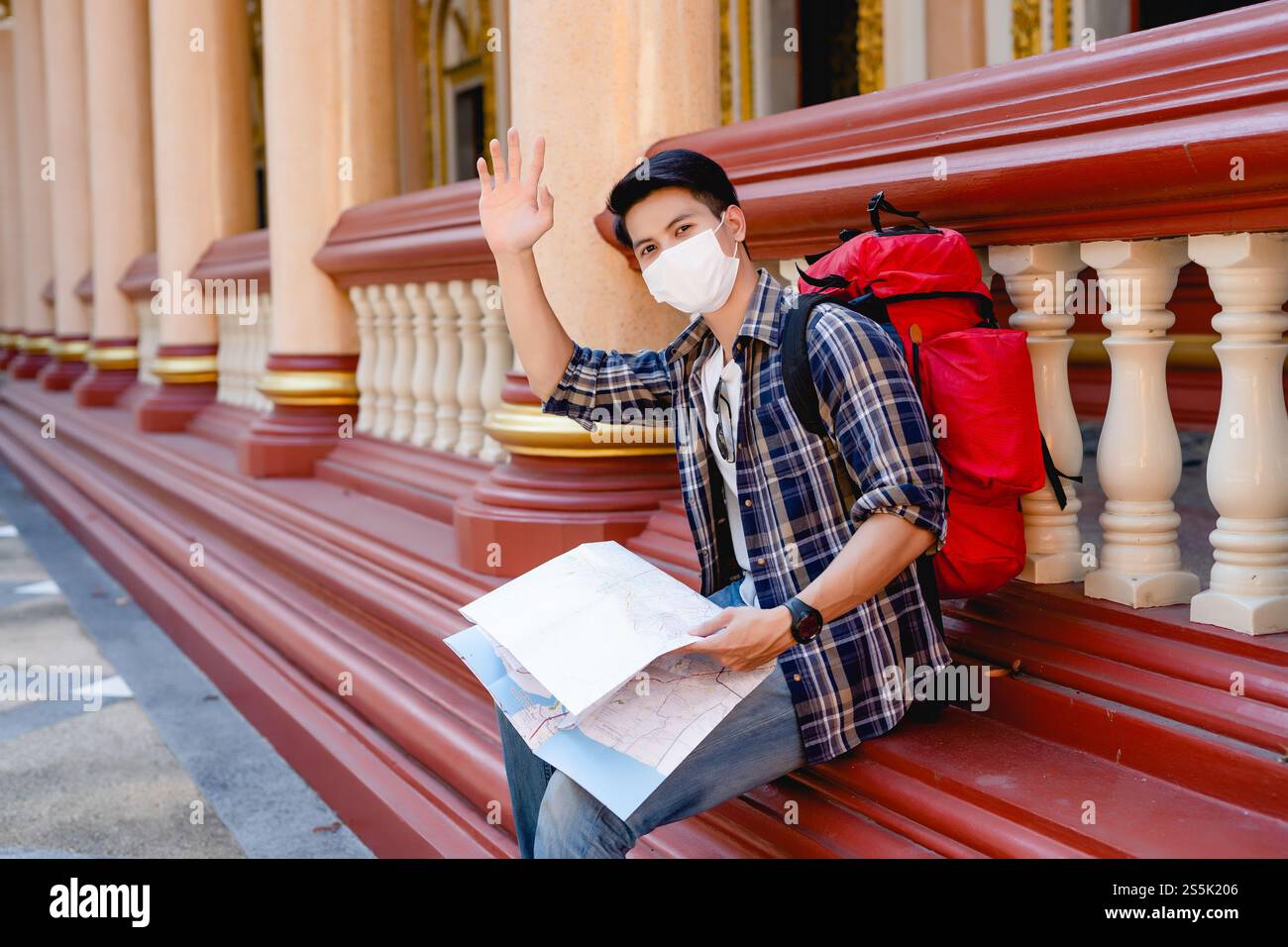Young Asian backpacker male in face mask sitting and hold a paper map ...