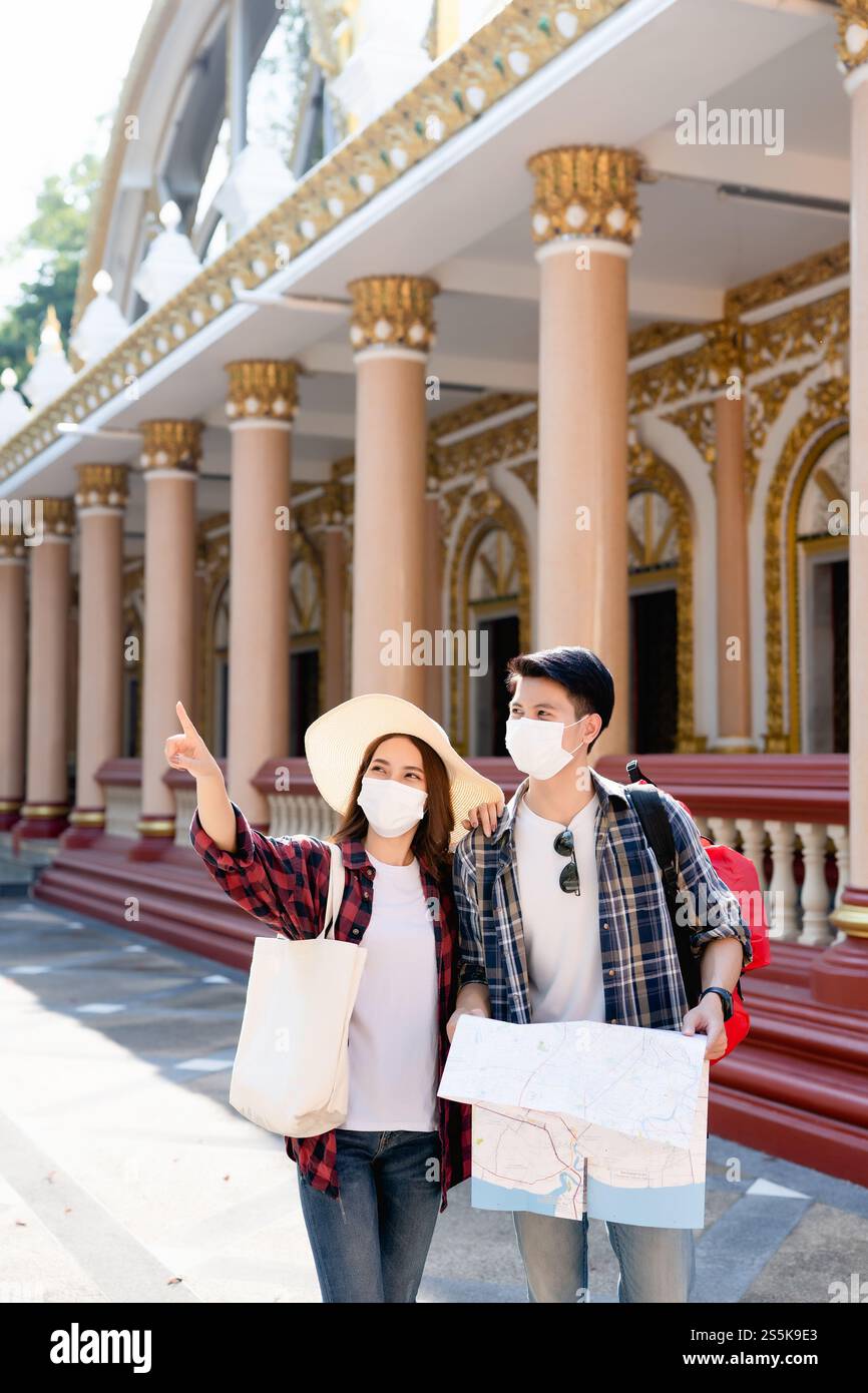 Young Asian backpacker couple in face mask at beautiful temple during vacations in Thailand, Pretty woman wearing sombrero, They are looking away Stock Photo