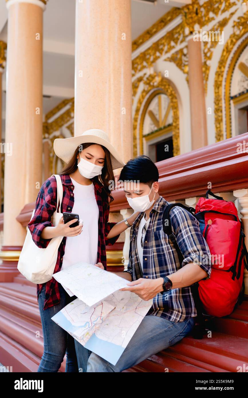 Young Asian backpacker couple in face mask at beautiful temple during vacations in Thailand, Pretty woman wearing sombrero, They use paper map and Stock Photo