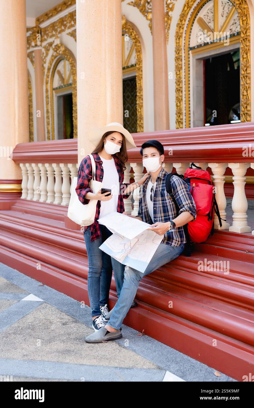 Young Asian backpacker couple in face mask at beautiful temple during vacations in Thailand, Pretty woman wearing sombrero, They use paper map and Stock Photo