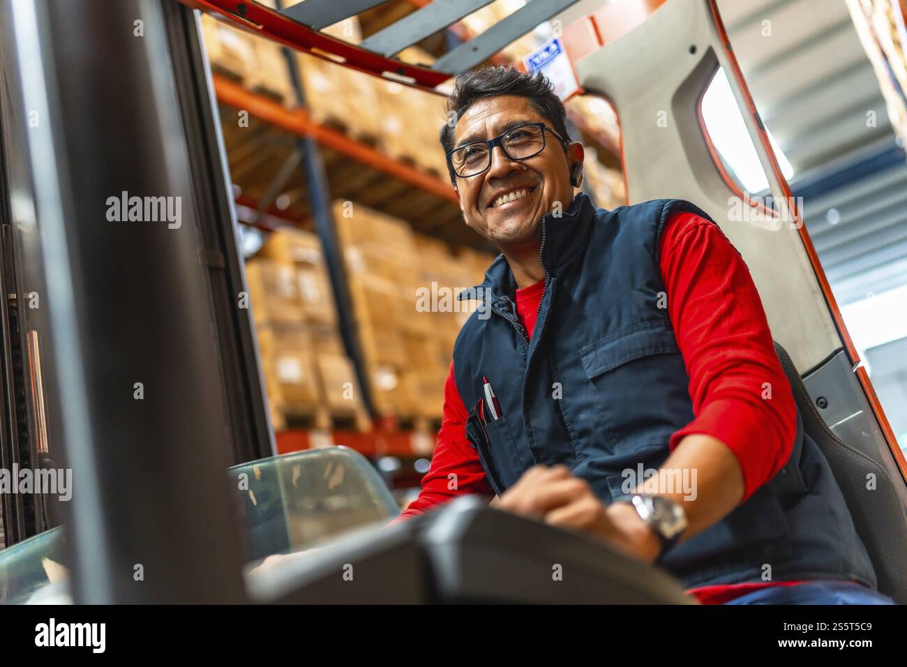 Happy latin worker smiling at camera while using forklift in a distribution warehouse Stock Photo
