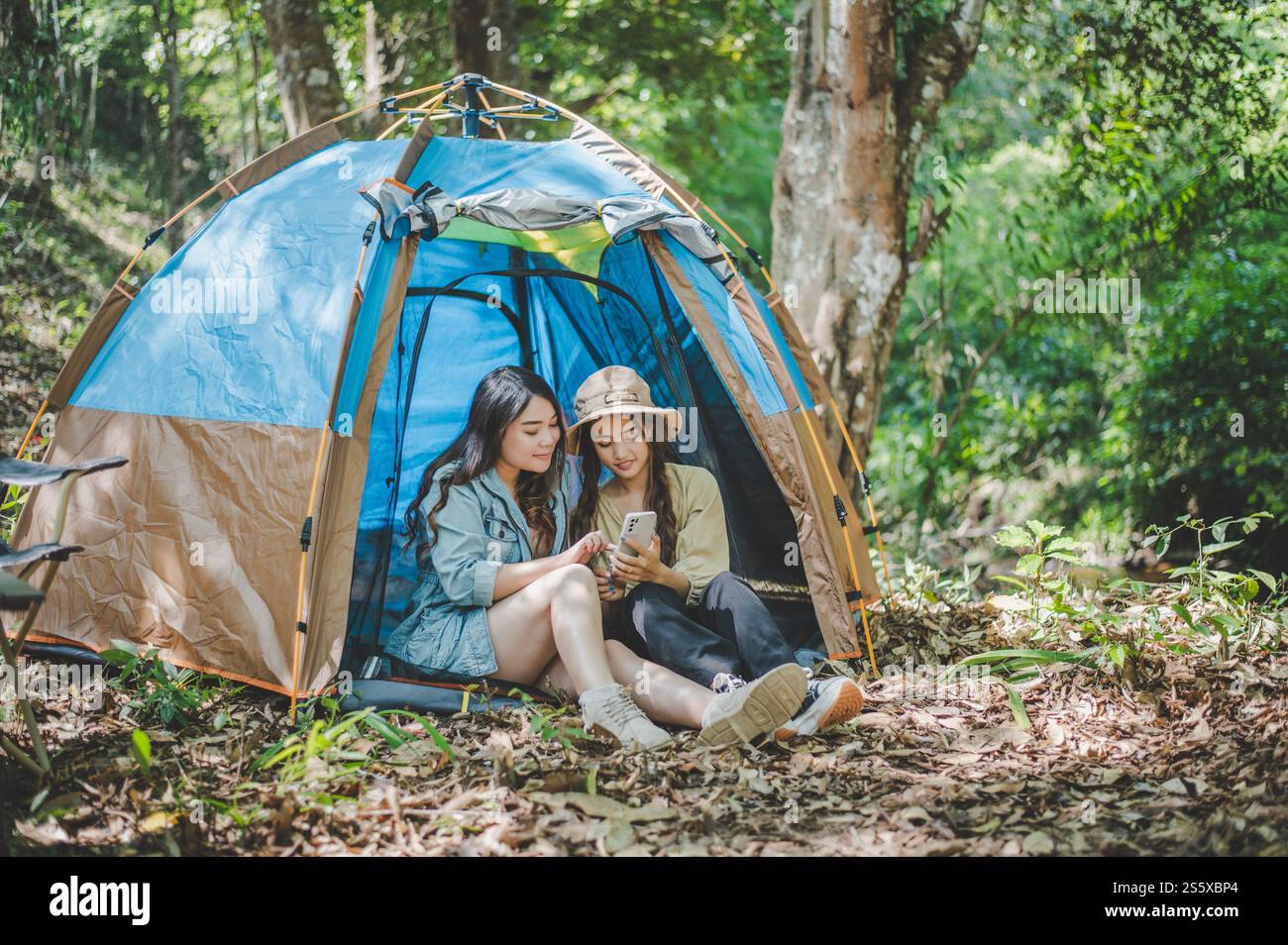 front view Young Asian pretty woman and her girlfriend sitting at front of tent, use mobile phone take photo during camping in forest with happiness Stock Photo