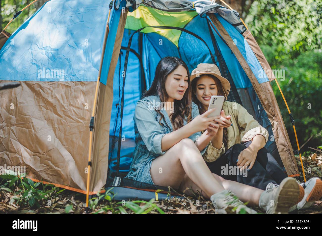 front view Young Asian pretty woman and her girlfriend sitting at front of tent, use mobile phone take photo during camping in forest with happiness Stock Photo