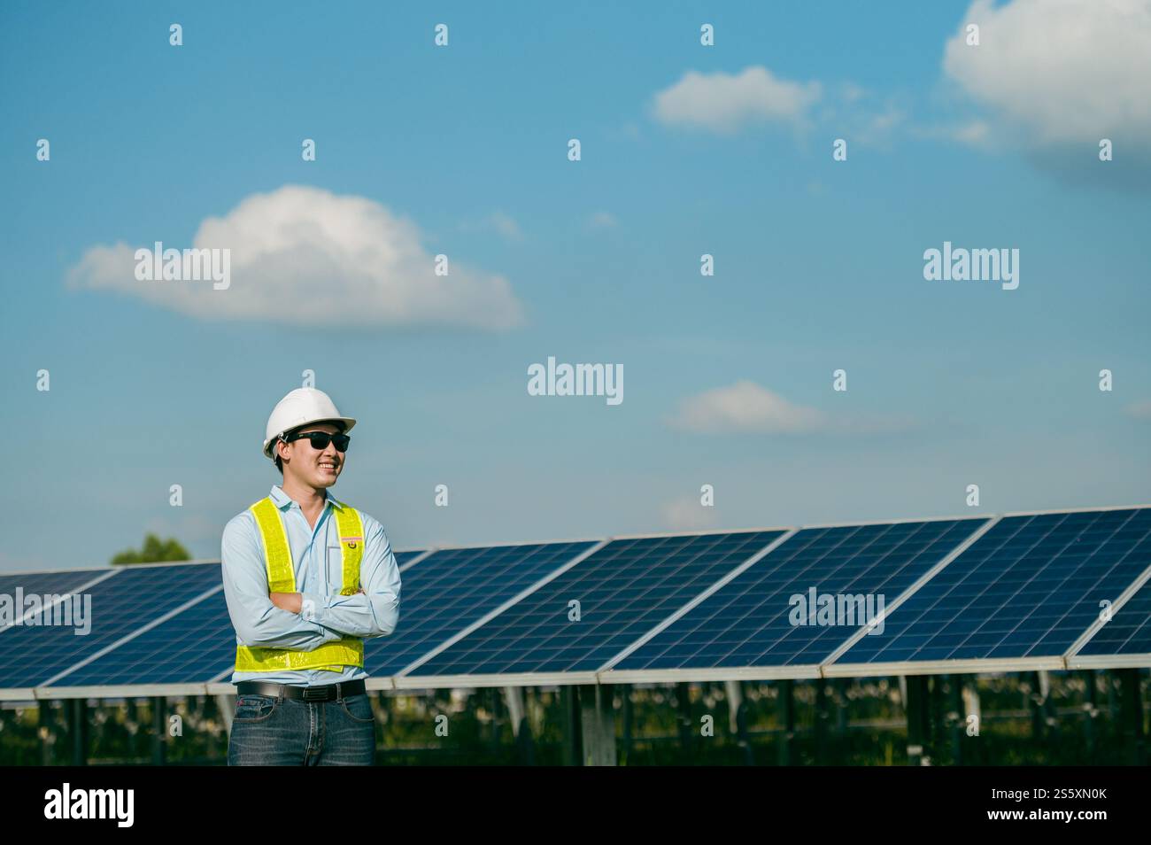 Portrait Asian Young engineer man wearing protective clothes, sunglasses and white helmet standing near solar panels while working in solar farm, Stock Photo