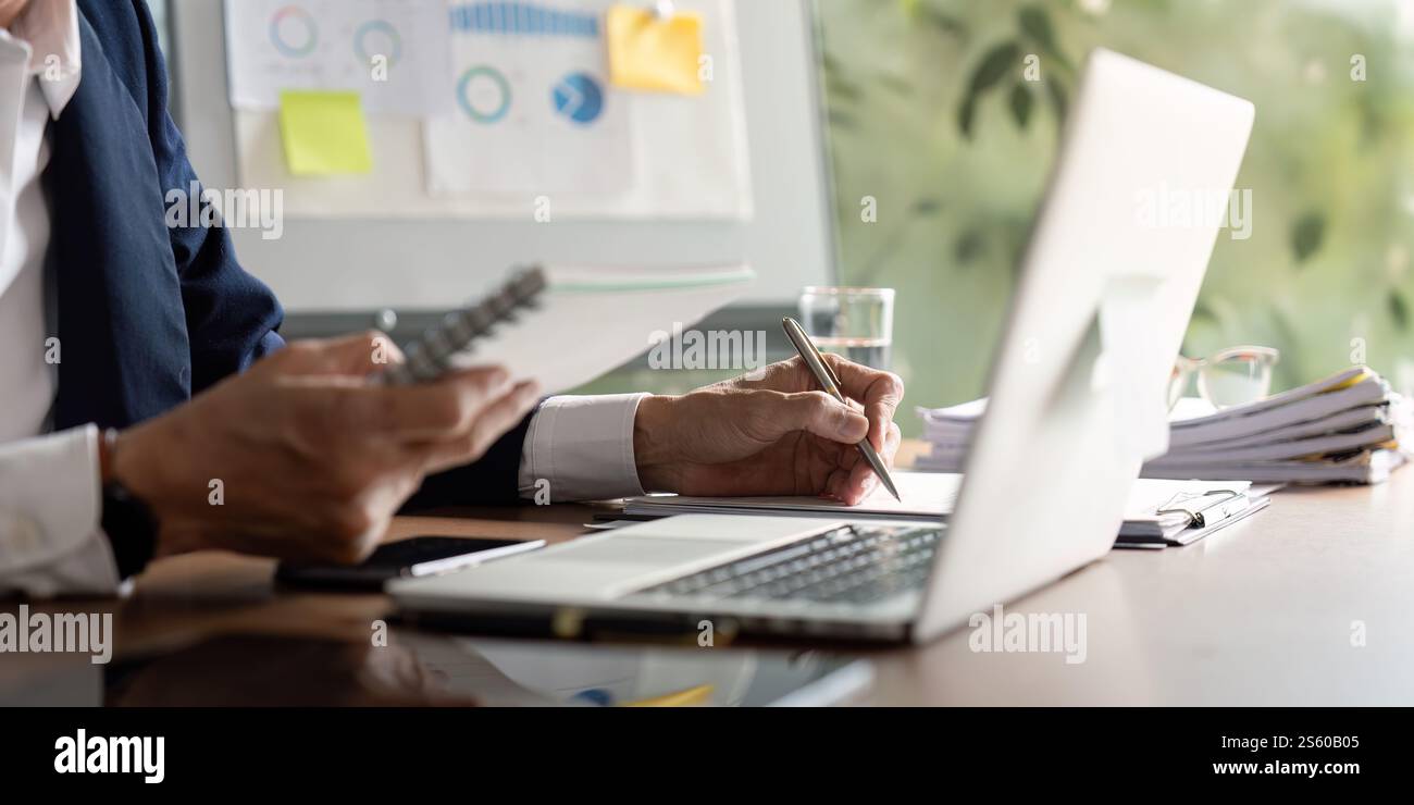 Elderly businessman taking notes while analyzing data on a laptop in a modern office Stock Photo