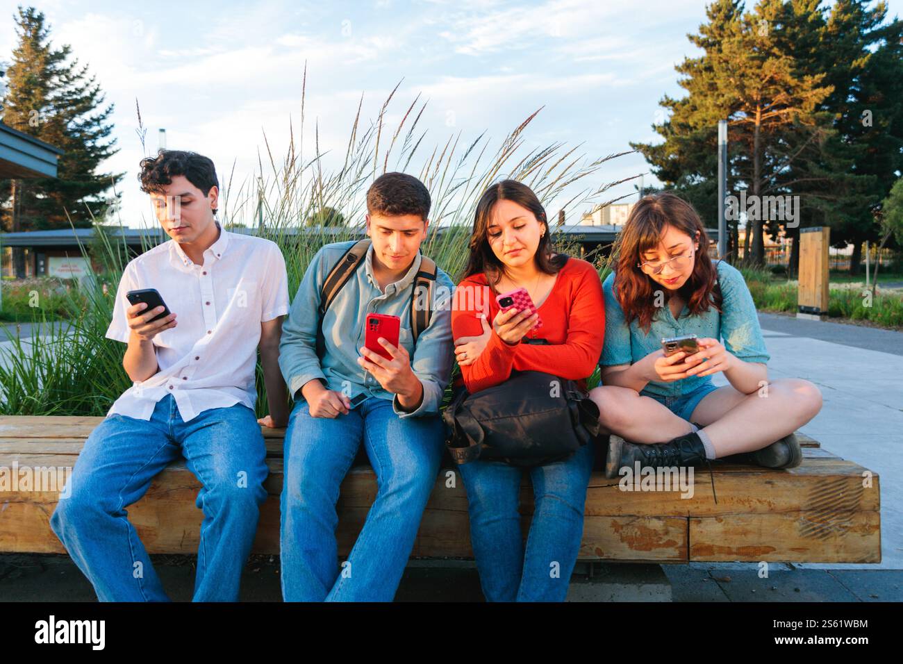 Young students using smartphones sitting on bench in university campus park Stock Photo