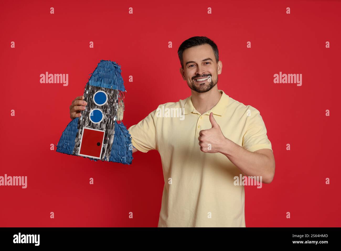 Happy man with rocket shaped pinata showing thumbs up on red background ...