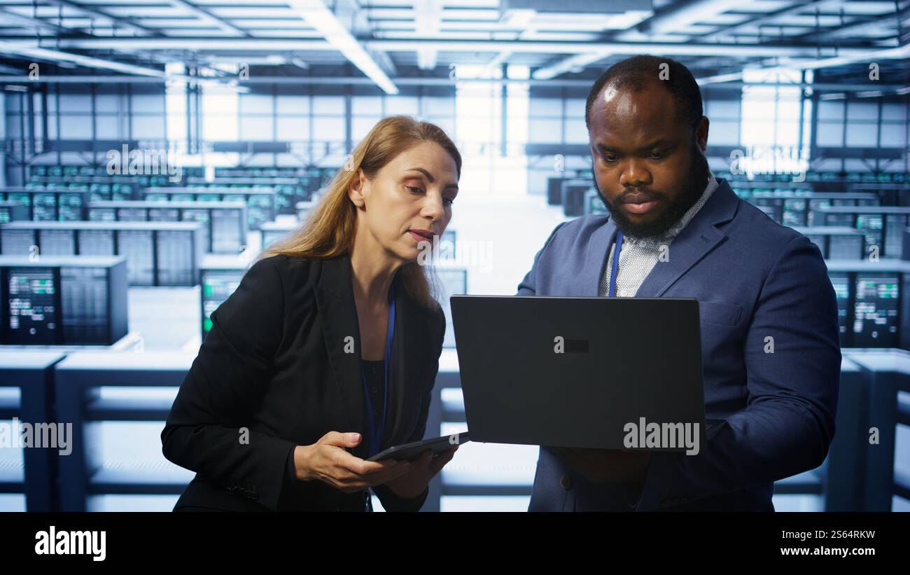 System administrators in data center overseeing server operations using notebook and tablet. Colleagues brainstorming in server hub, ensuring optimal performance and data integrity, camera B Stock Photo