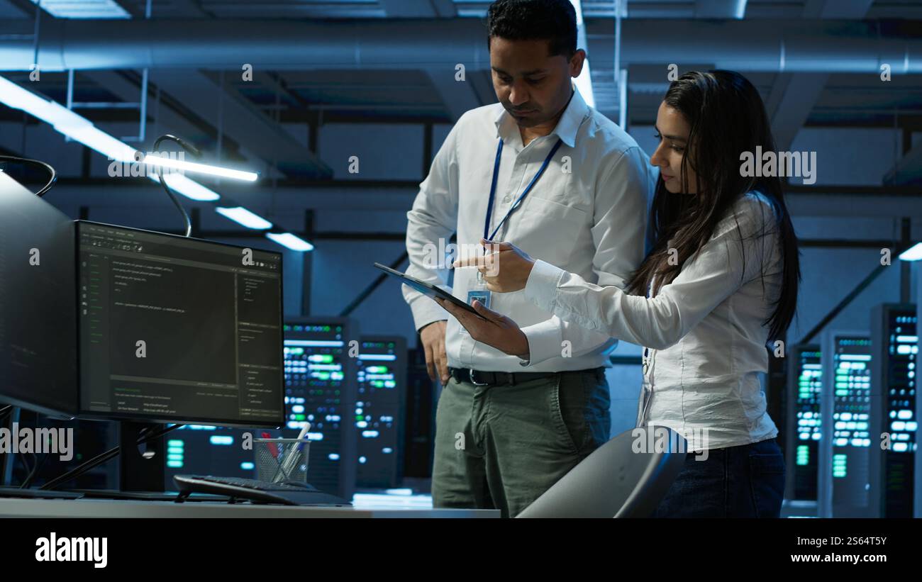 Team of programmers in high tech data center troubleshooting issues using tablet. Teamworking colleagues in server farm using device, establishing performance benchmarks, camera A Stock Photo