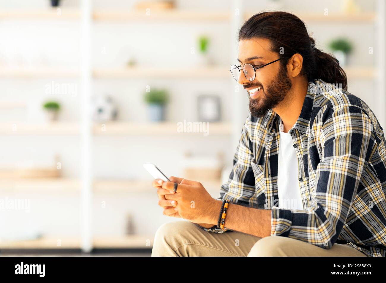 Positive long-haired middle-eastern guy sitting on couch, using cellphone Stock Photo