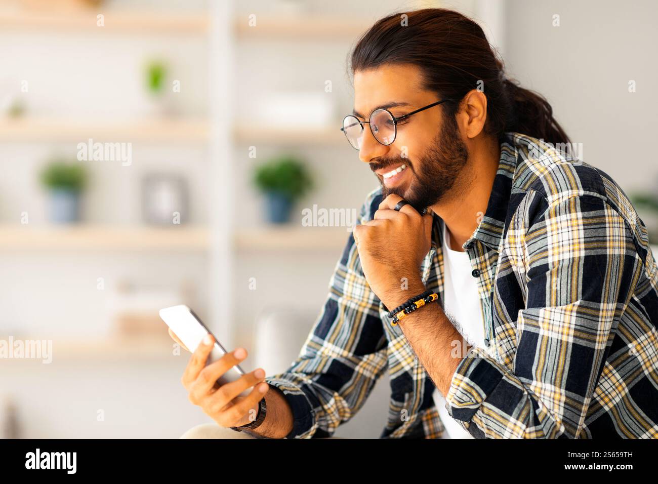 Cheerful long-haired middle-eastern guy hipster sitting on couch, using smartphone Stock Photo