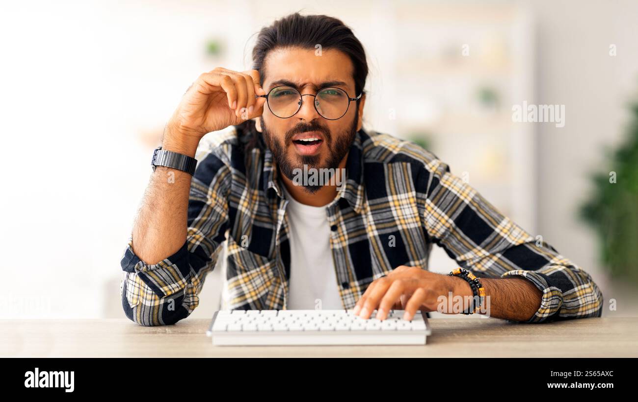 Confused middle-eastern man programmer sitting at worktable, touching glasses Stock Photo