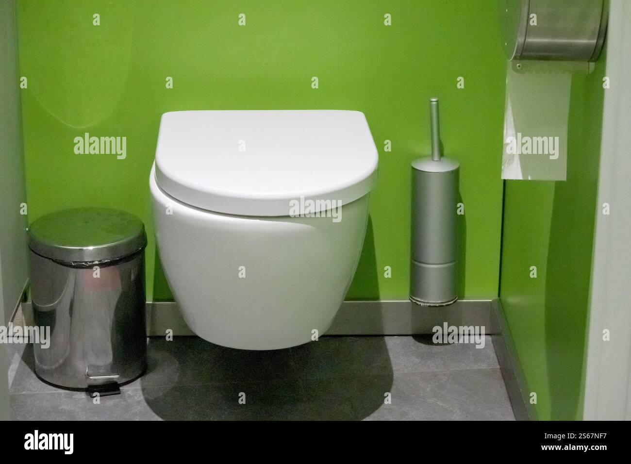 A white wall-mounted toilet bowl with the lid down, a silver toilet brush and a metal trash can on a bright green wall background. Stock Photo