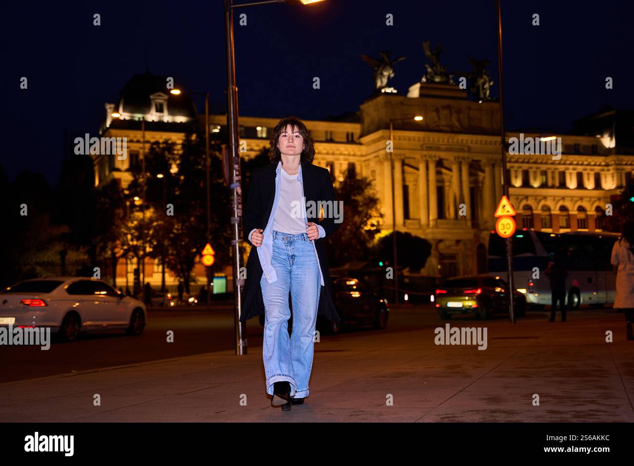 Stylish young woman walking confidently on a city street at night, with ...