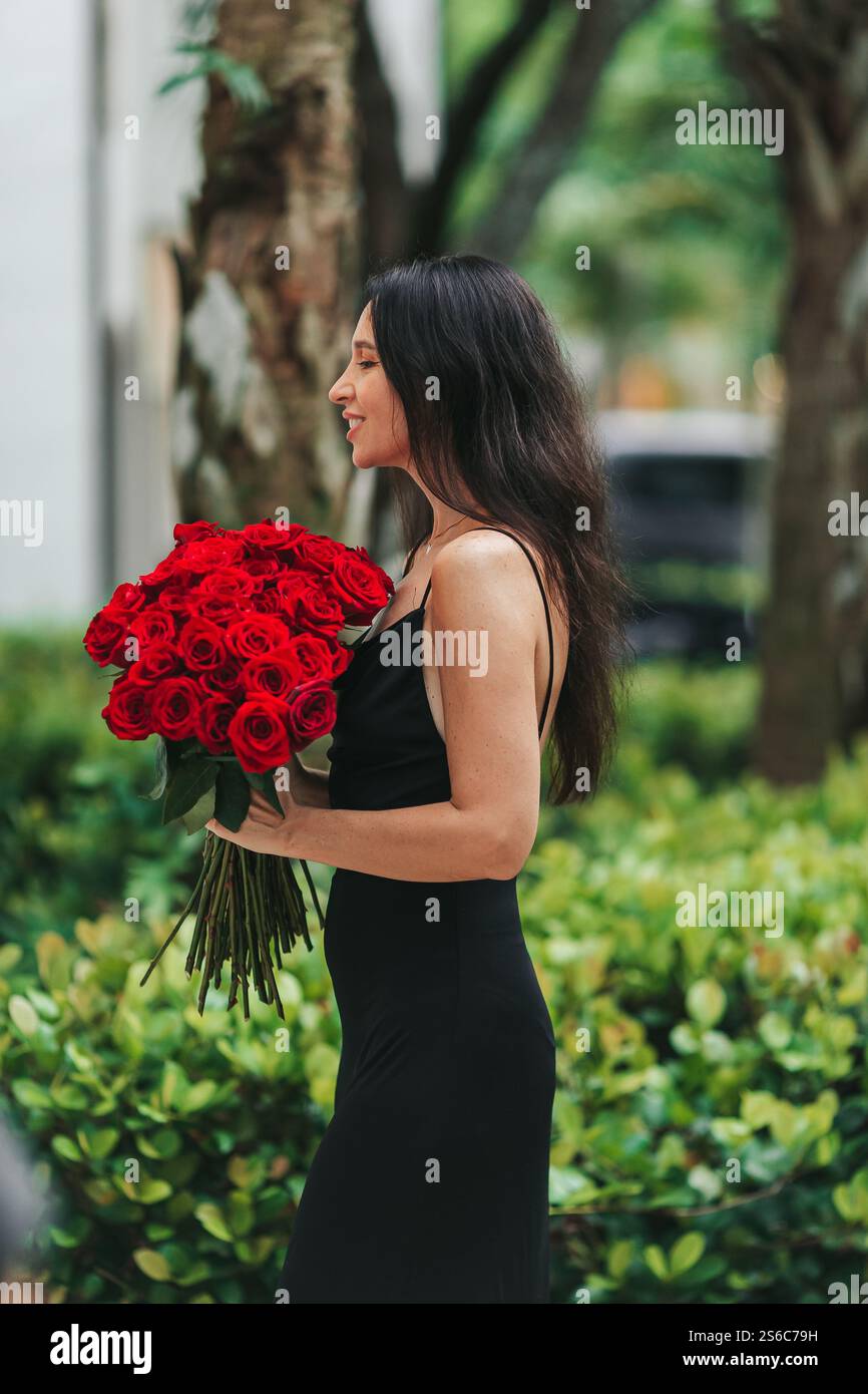 Happy young woman in black dress holding bouquet of red roses outdoors ...