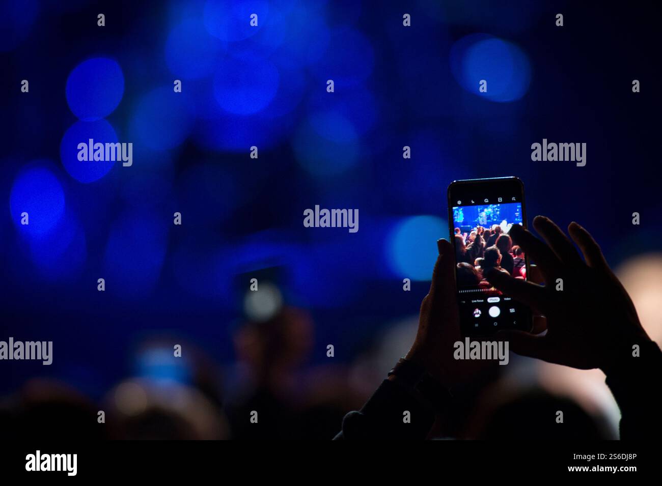 A smartphone frames the concert scene, capturing a fleeting moment amidst the crowd. The blue bokeh adds atmosphere, balancing connection & immersion. Stock Photo