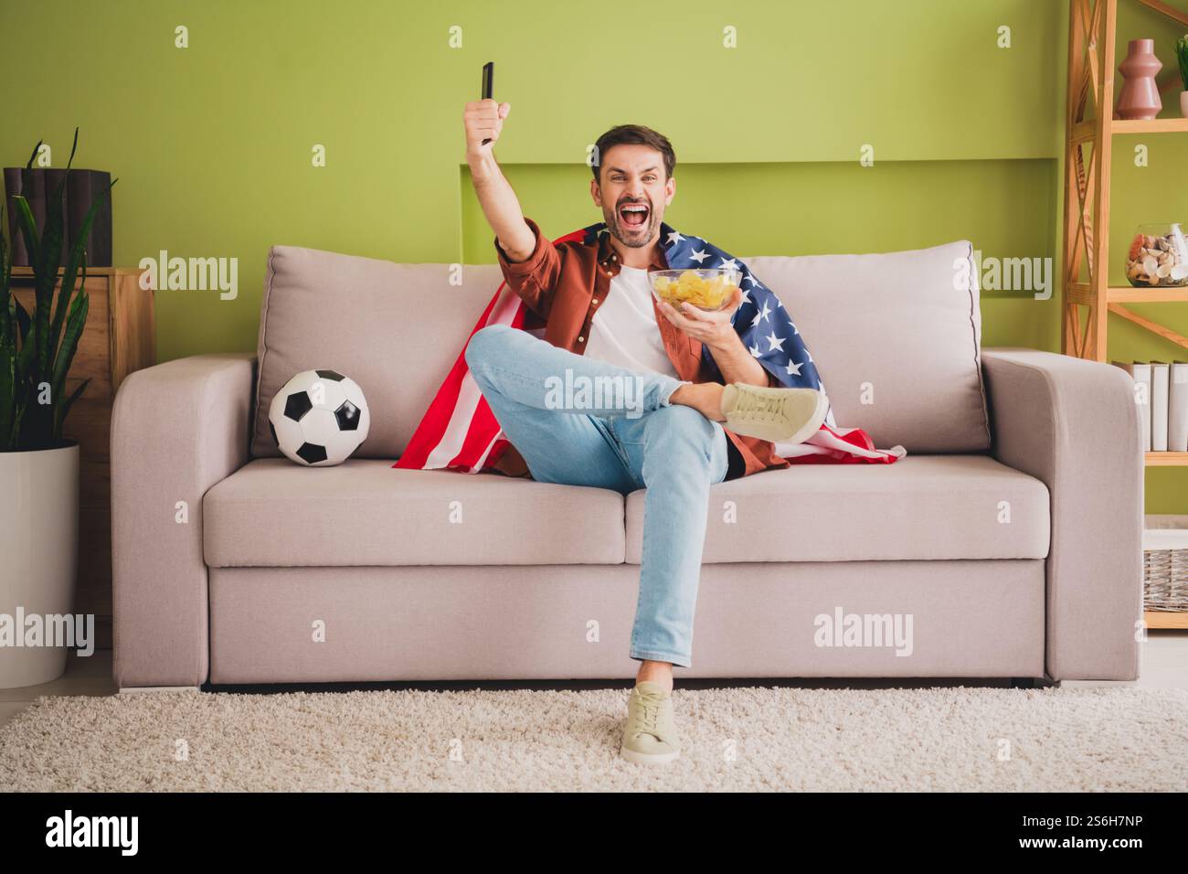Excited young soccer fan indoors enjoying a game with snacks and casual ...