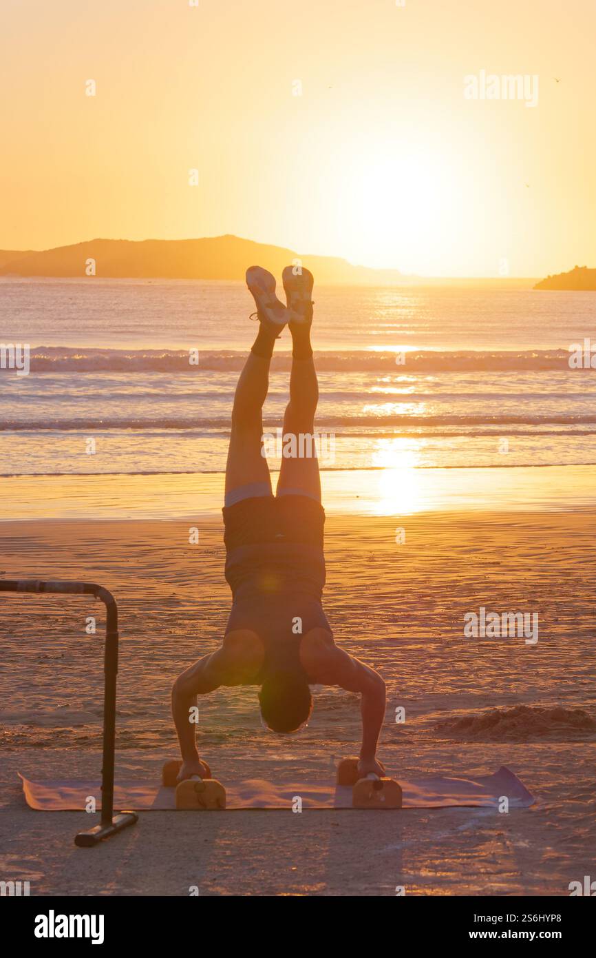 Athletic young man performs a hand stand on the beach as waves roll in at sunset with Island behind. Essaouira, Morocco. Jan, 2025 Stock Photo