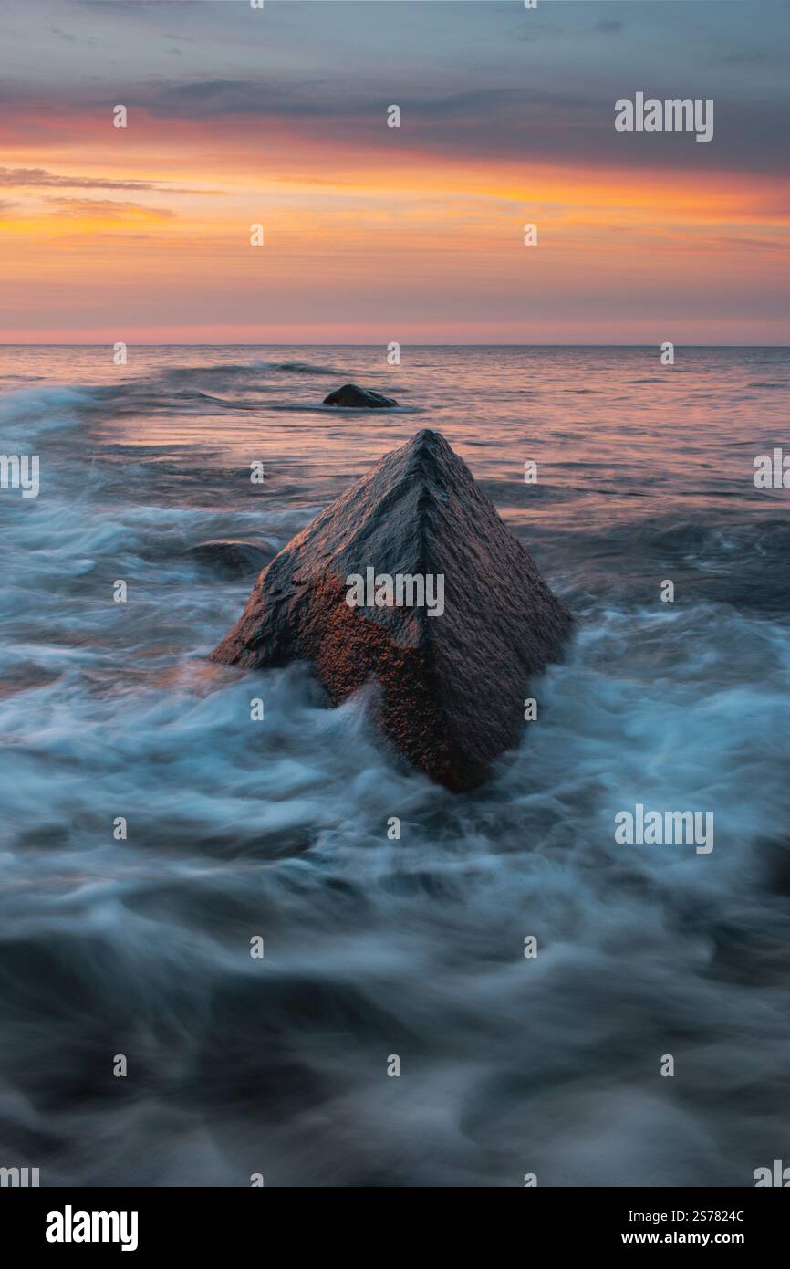 A long exposure shot of Montauk, NY, at sunset with soft waves crashing against a triangular rock, creating a serene and dynamic ocean scene. Stock Photo