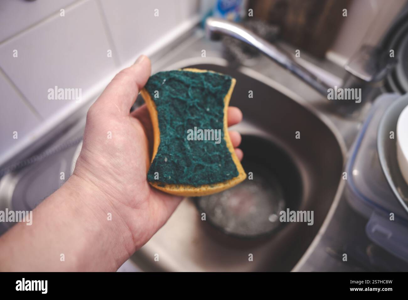 Hand holding a green kitchen sponge over a sink full of soapy water ...