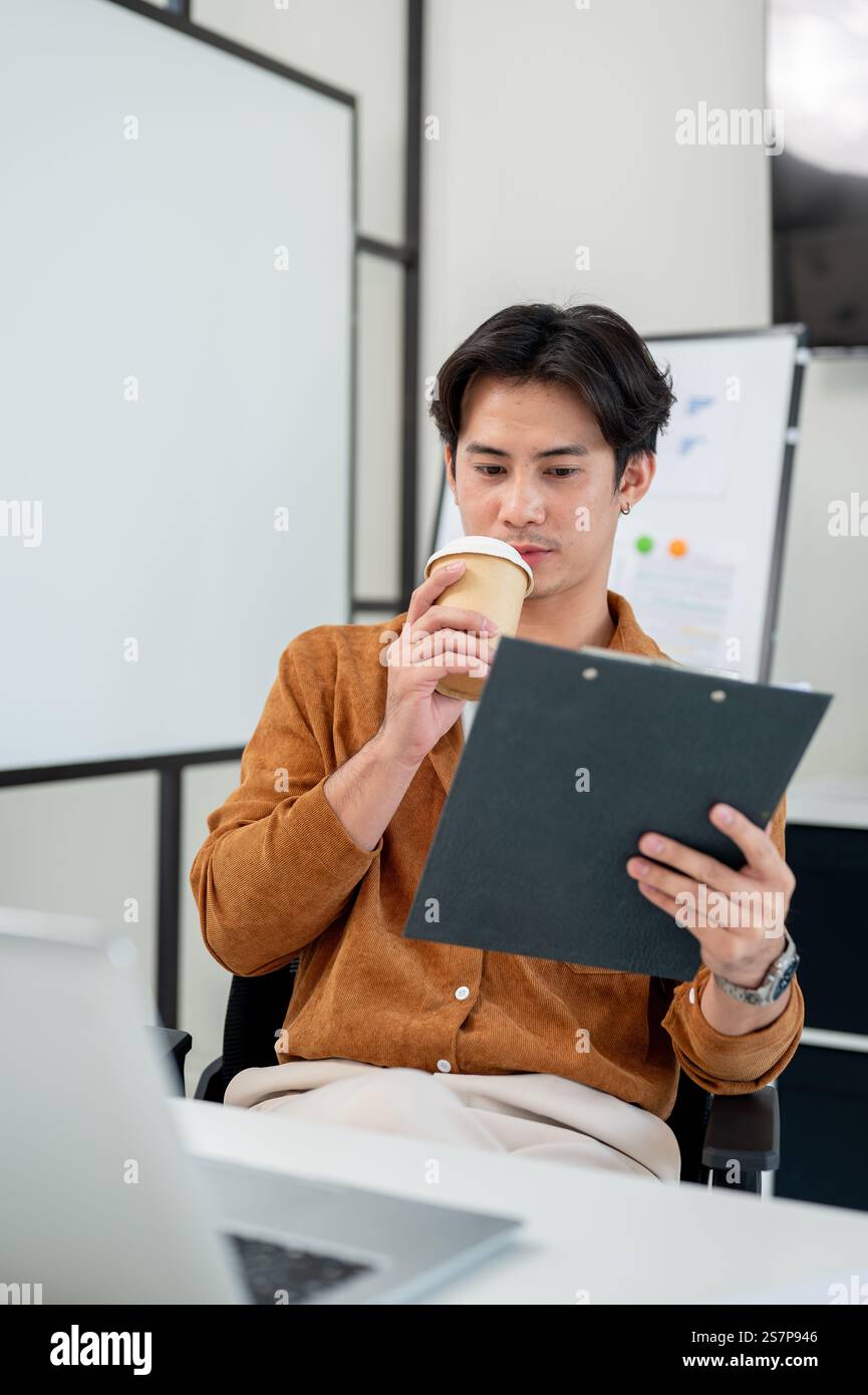 A smart, handsome Asian male office worker focuses on reading a document at his desk in the office. corporate job, marketing agency, businessman Stock Photo