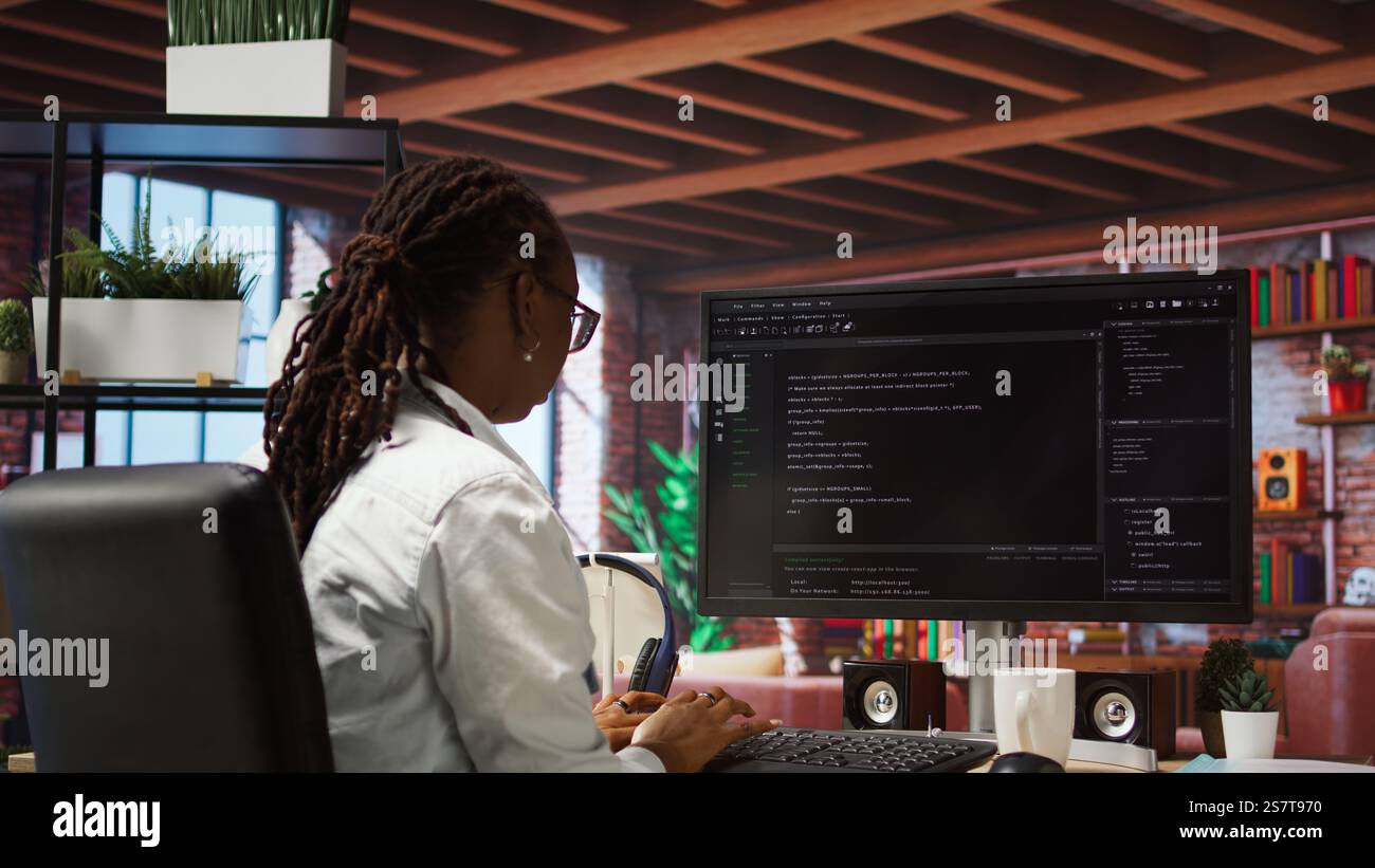 Programmer types code into PC, entering commands, running script. African american IT professional in apartment fixing errors in application, implementing new features, camera A Stock Photo