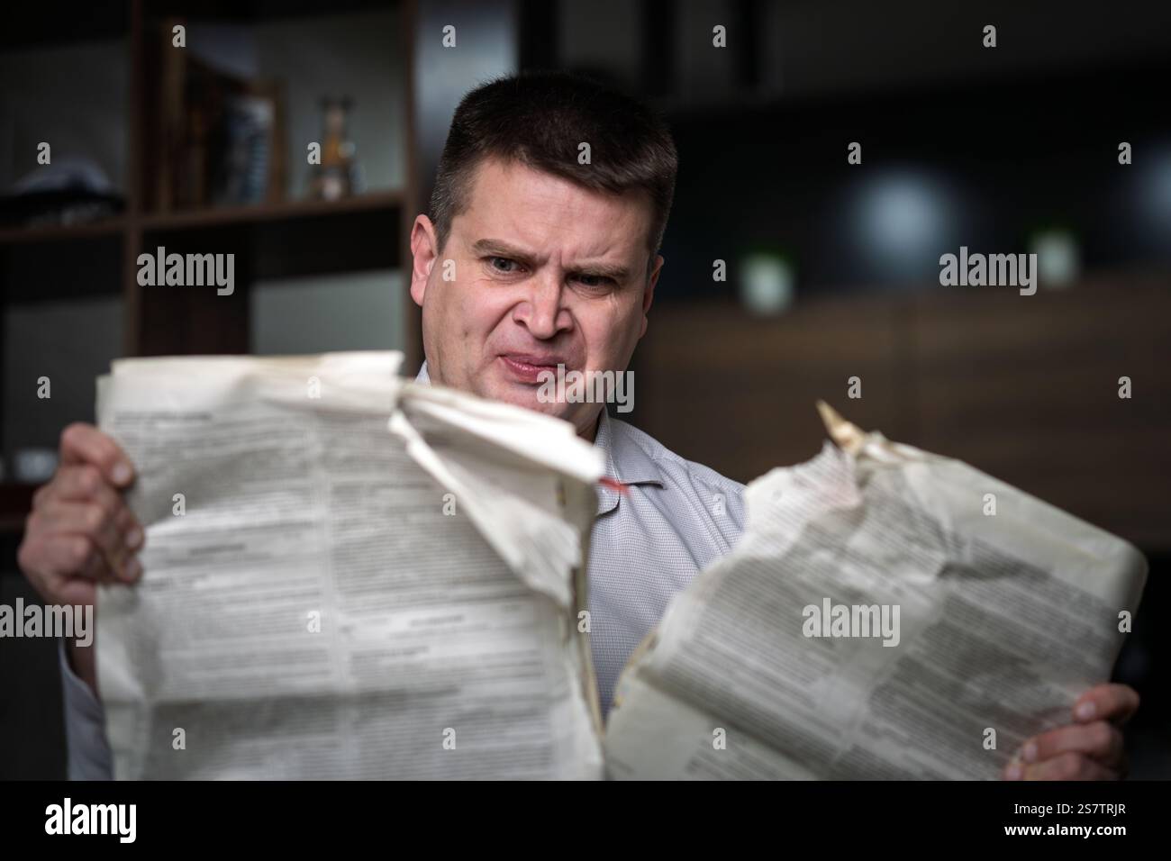 A man in a white shirt reads a newspaper at home against a dark background. concept of deception in the press. a man tears up a newspaper with false n Stock Photo