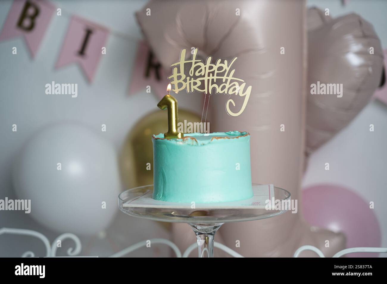 A teal birthday cake with gold accents, topped with a lit golden number 1 candle and a 'Happy Birthday' topper. The cake sits on a glass stand against Stock Photo