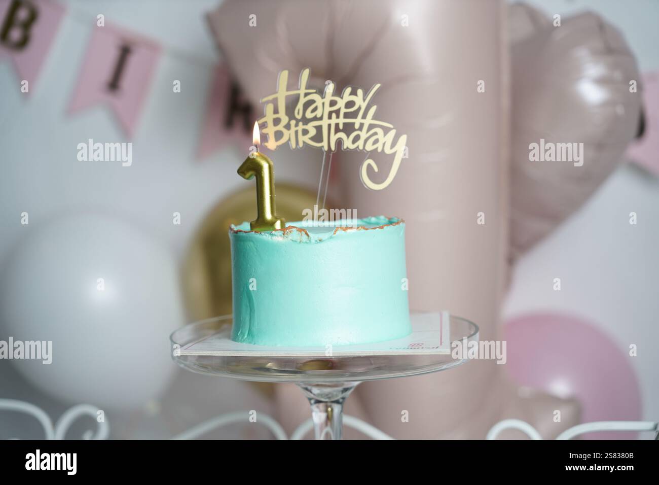 A teal birthday cake with gold accents, topped with a lit golden number 1 candle and a 'Happy Birthday' topper. The cake sits on a glass stand against Stock Photo