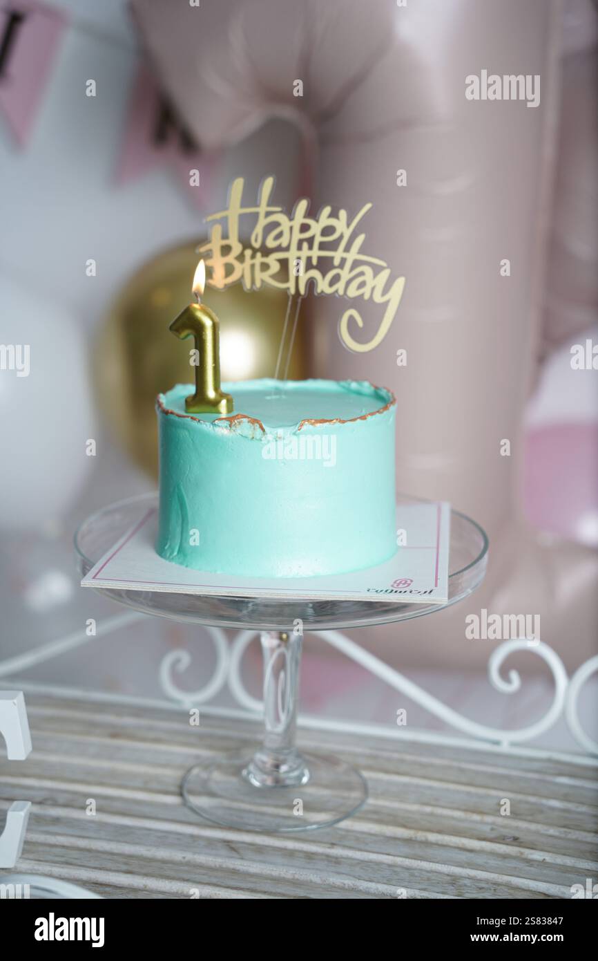 A close-up of a teal birthday cake with gold accents, topped with a lit golden number 1 candle and a 'Happy Birthday' topper. The blurred background f Stock Photo