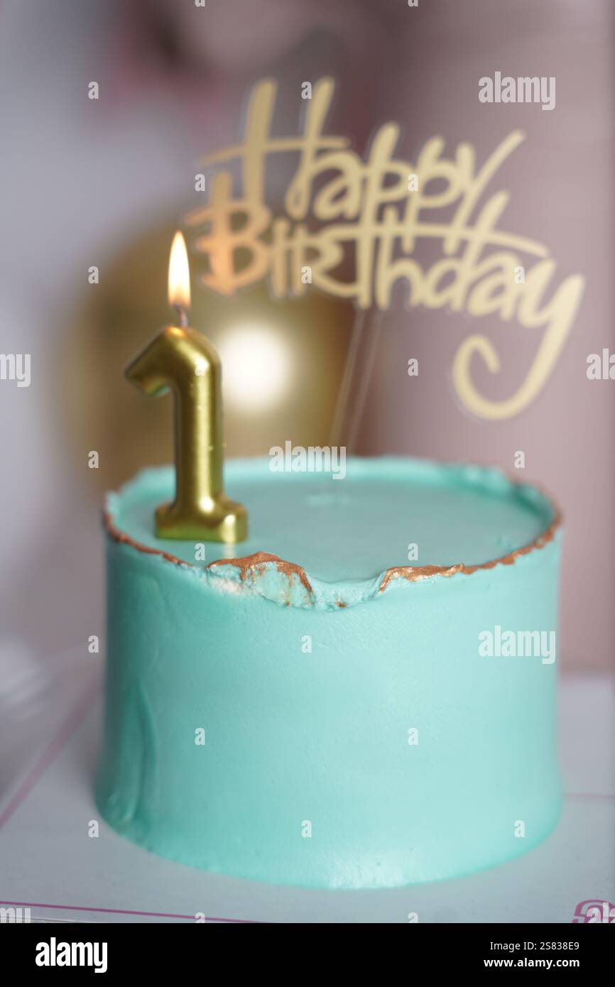 A close-up of a teal birthday cake with gold accents, topped with a lit golden number 1 candle and a 'Happy Birthday' topper. The blurred background f Stock Photo