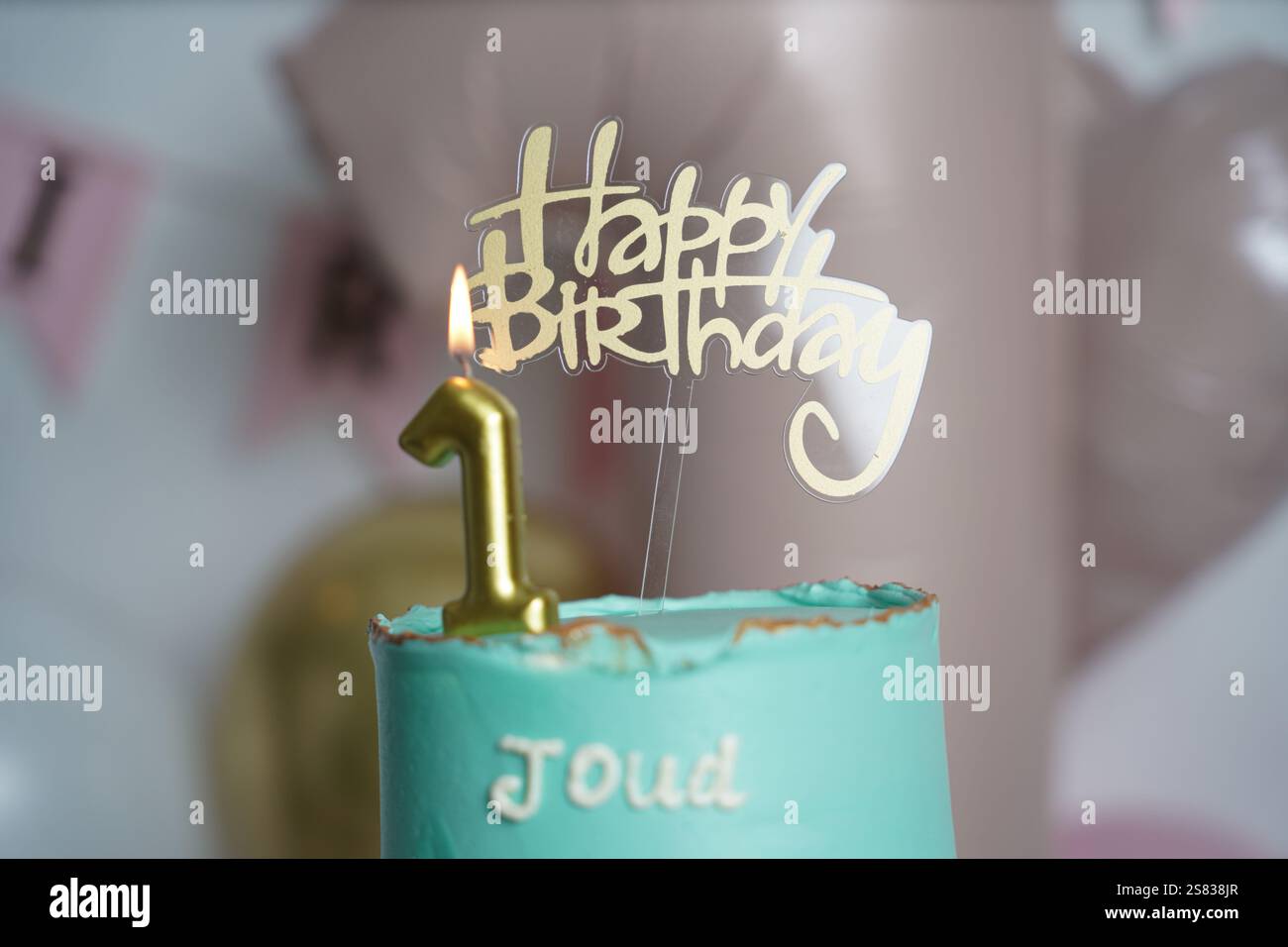 A teal birthday cake with gold accents, topped with a lit golden number 1 candle and a 'Happy Birthday' topper. The cake sits on a glass stand against Stock Photo