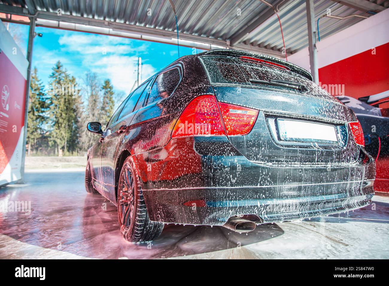 A black station wagon covered in soap suds is parked under a metal roof at a car wash. Tall evergreen trees and a clear blue sky are visible in the ba Stock Photo