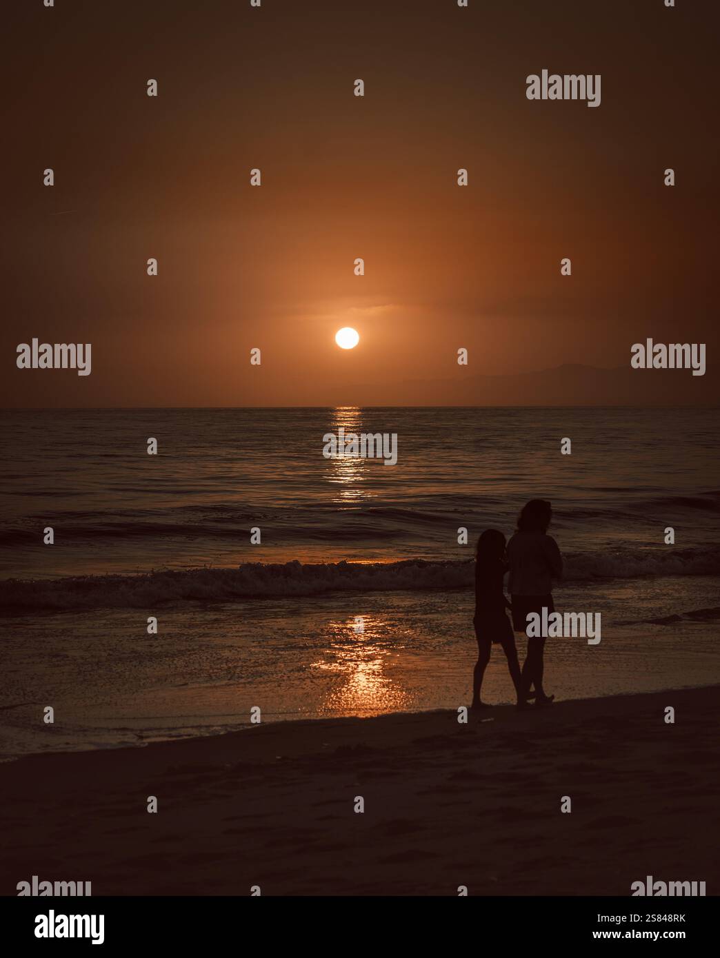 Silhouettes of two people walking along the beach during a serene sunset, with the sun reflecting on the calm ocean waves. Stock Photo