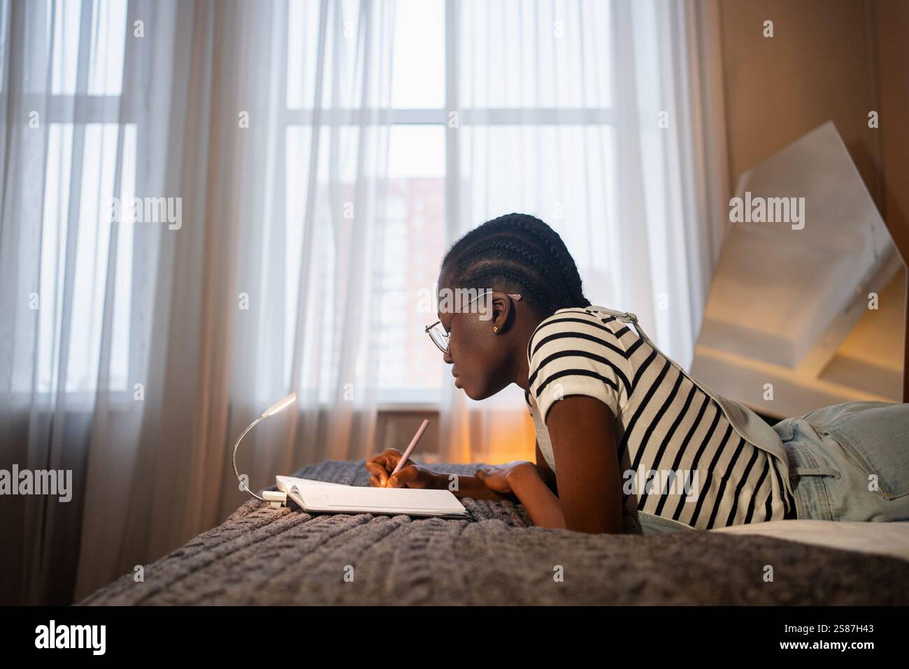 Focused young black woman wearing glasses is lying on her stomach on her bed, diligently writing in a notebook under the warm glow of a small lamp Stock Photo