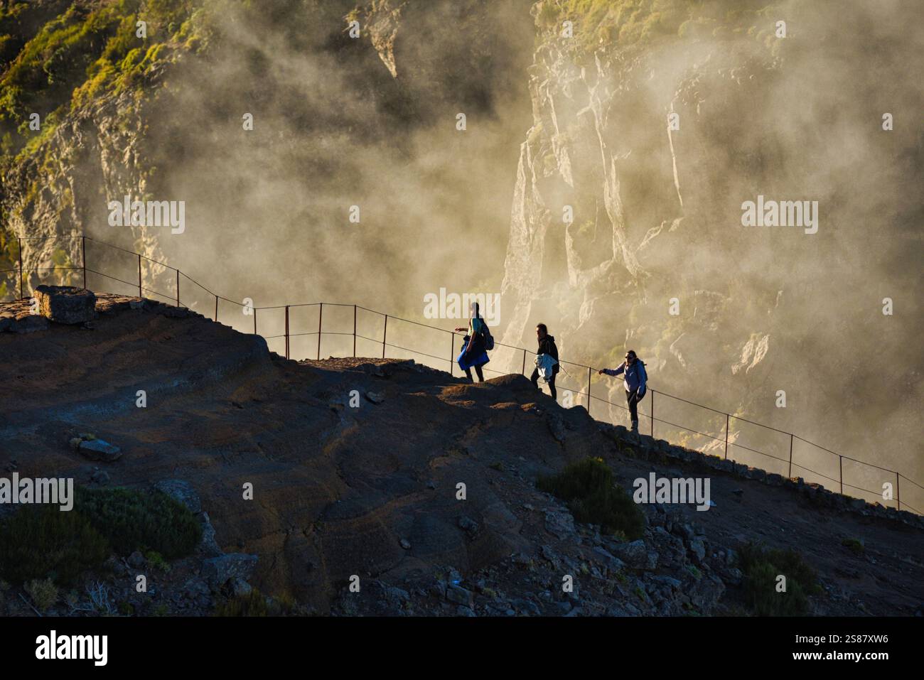 Hikers on trail pathway along jagged volcanic cliffs on Pico Areeiro with cloud inversions blowing over mountain peaks, Madeira island, Portugal. Stock Photo