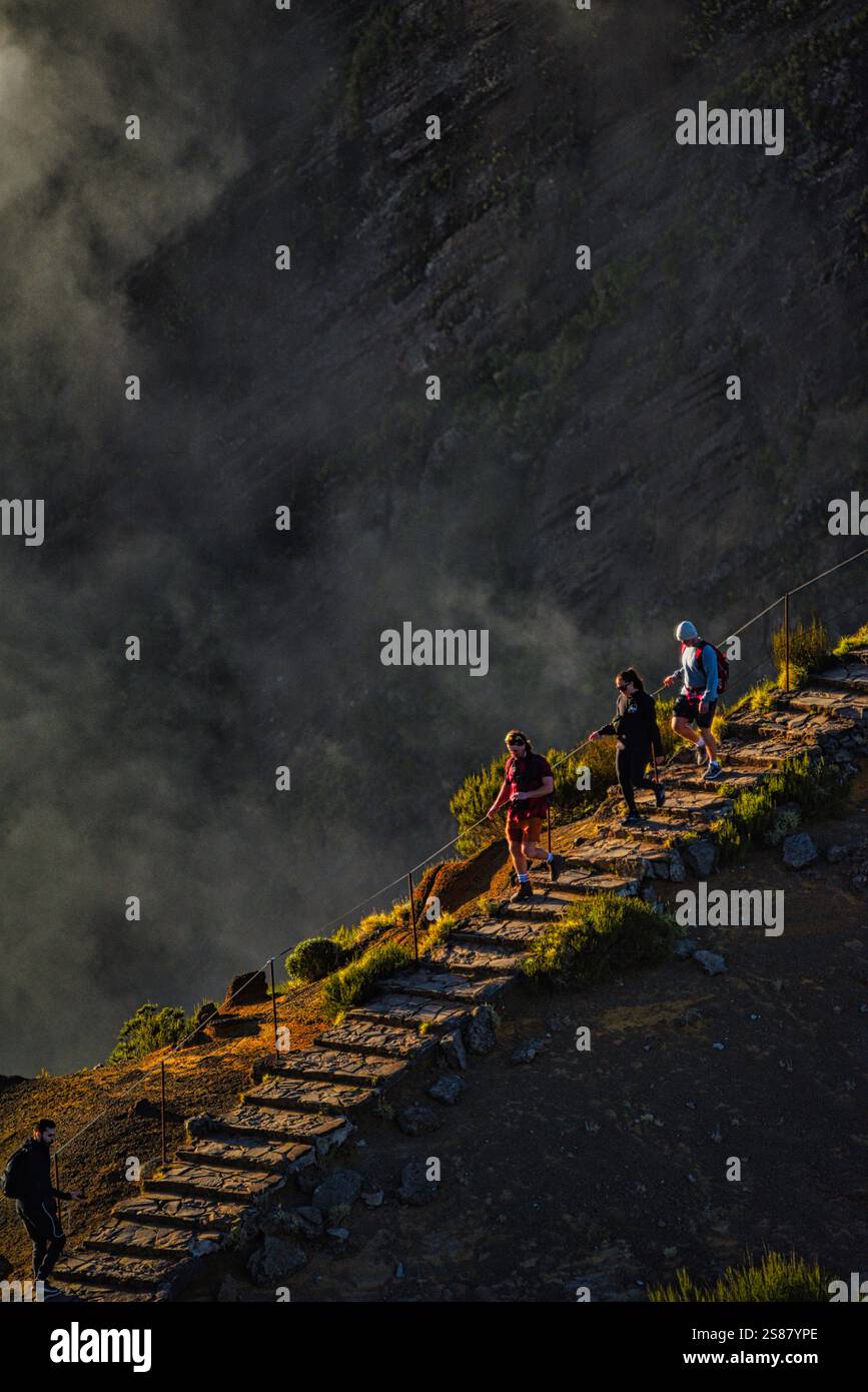 Hikers on trail pathway along jagged volcanic cliffs on Pico Areeiro with cloud inversions blowing over mountain peaks, Madeira island, Portugal. Stock Photo