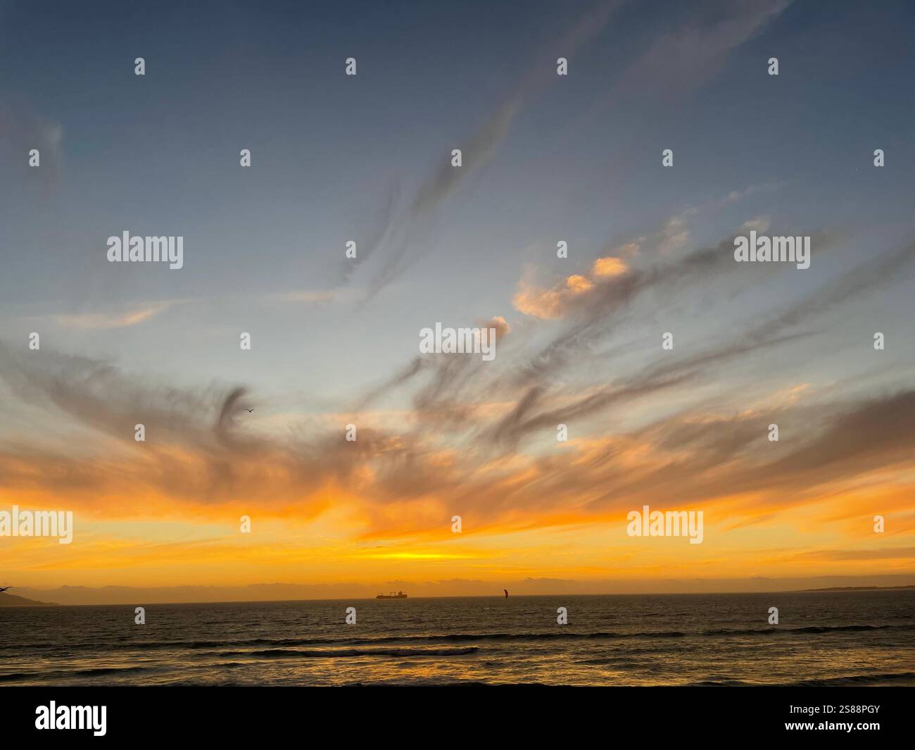 sun going down over the horizon and sea with a ship in the distance and streaky grey clouds in a blue orange sky at sunset in Cape Town, South Africa - Smartphone Captured Stock Image