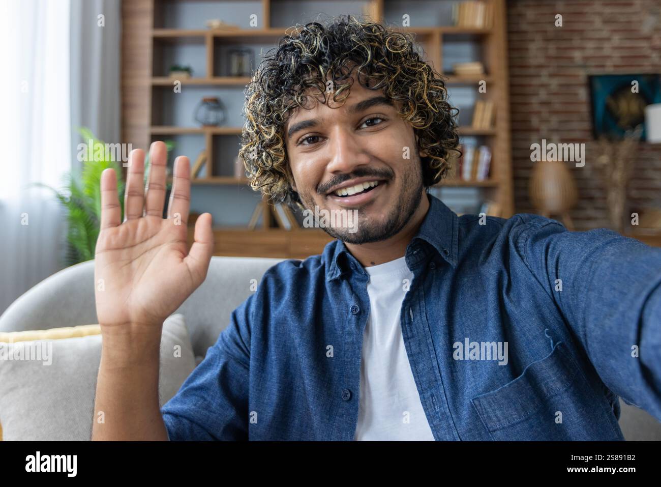 A man looks into the phone camera, happily waves his hand, takes a selfie photo and talks to colleagues and friends remotely using a smartphone app for video chats. Stock Photo