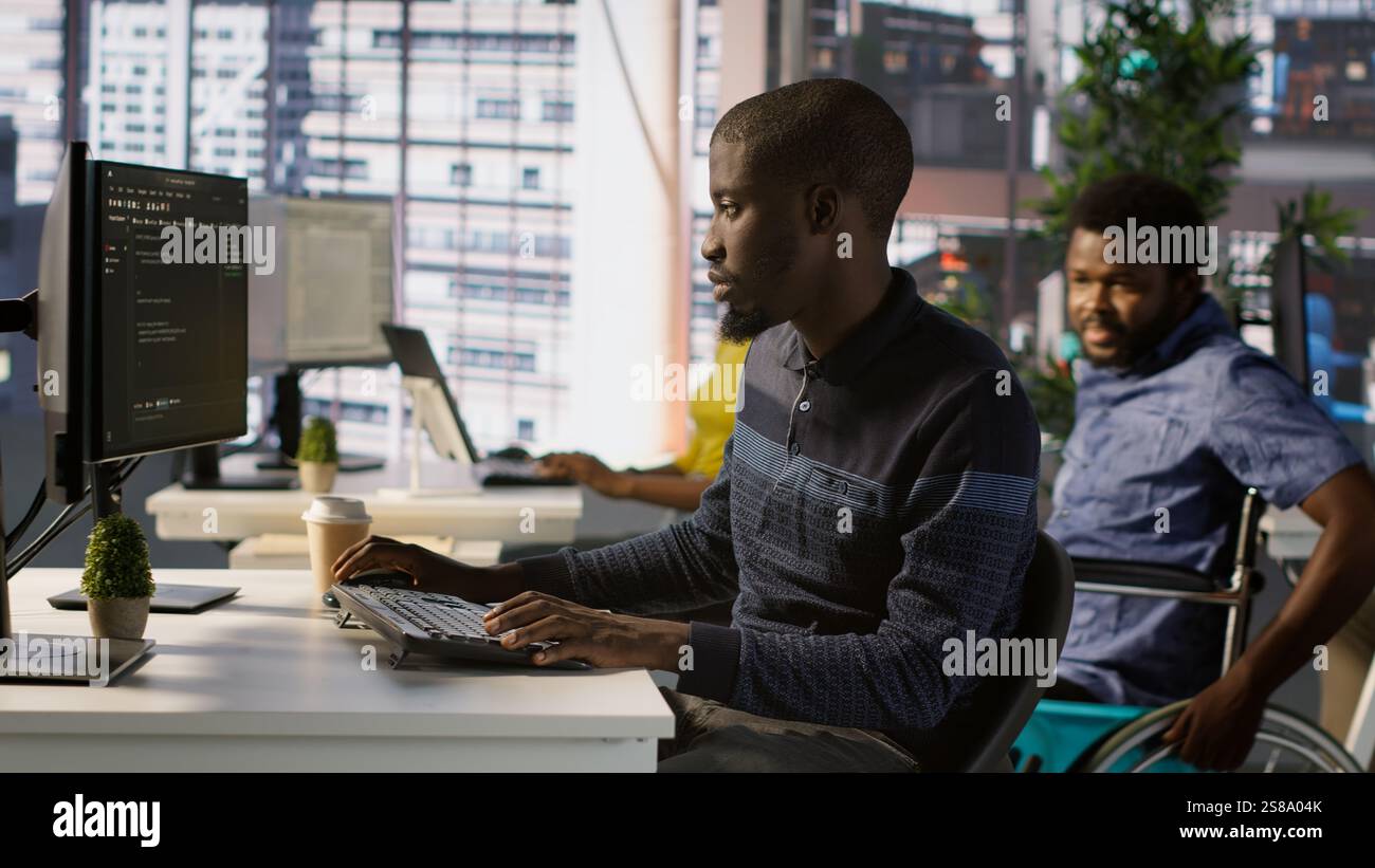 Programmer doing software debugging, asking for help from smiling team leader in wheelchair. Employee fixing coding mistakes, being assisted by cheerful manager with disability, camera A Stock Photo