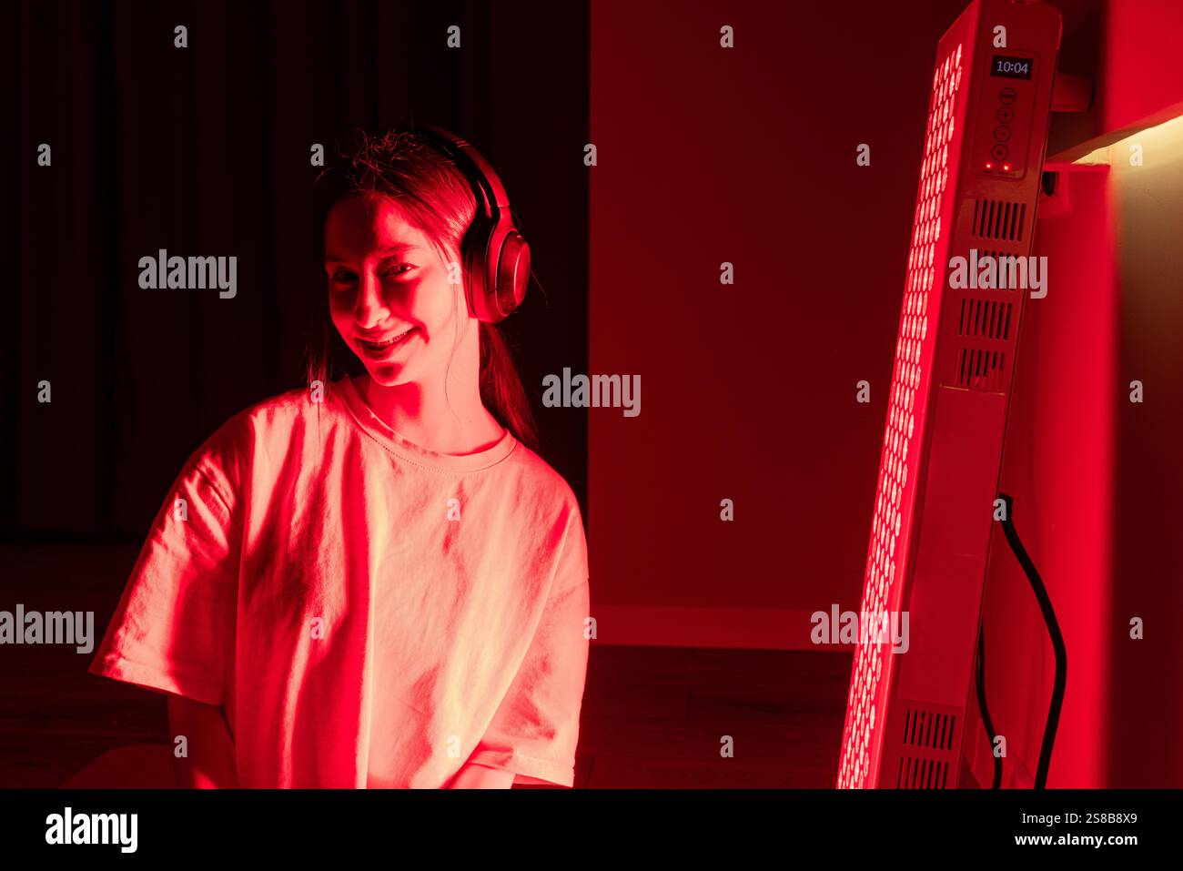 A young woman smiling indoors, wearing headphones, during red light therapy Stock Photo