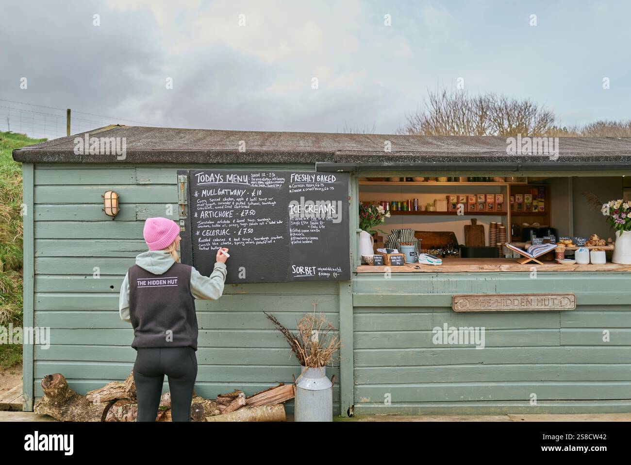 A waitress chalks up the winter menu soups at the Hidden Hut cafe at Porthcurnick Beach, Cornwall, UK Stock Photo