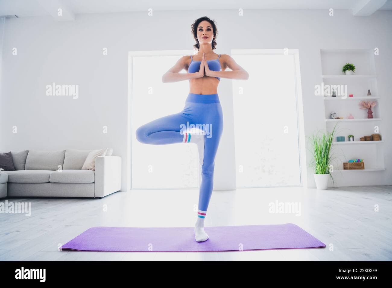 Young woman practicing yoga at home, balancing in tree pose in bright ...