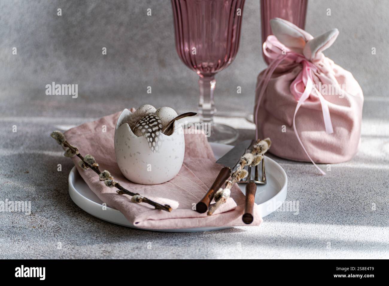 Beautifully arranged Easter table setting featuring a cracked eggshell vase with feathers, surrounded by pink elements and spring decorations. Soft li Stock Photo